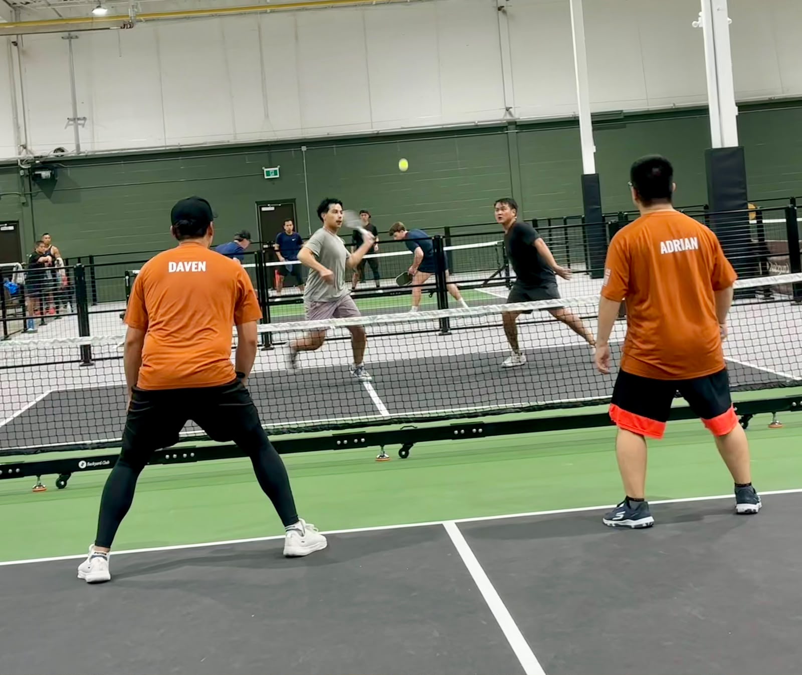 Daven and Adrian from behind mid-rally, names on the backs of their orange shirts, as the ball floats over to the other pair