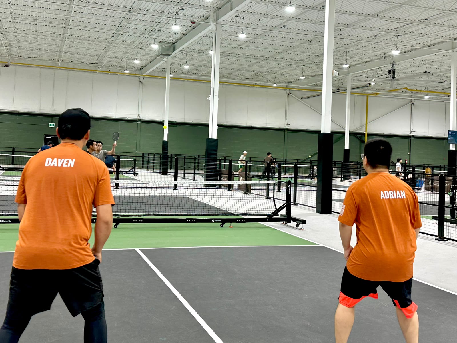 Daven and Adrian from behind on the baseline, names across their orange shirts, waiting on the next shot