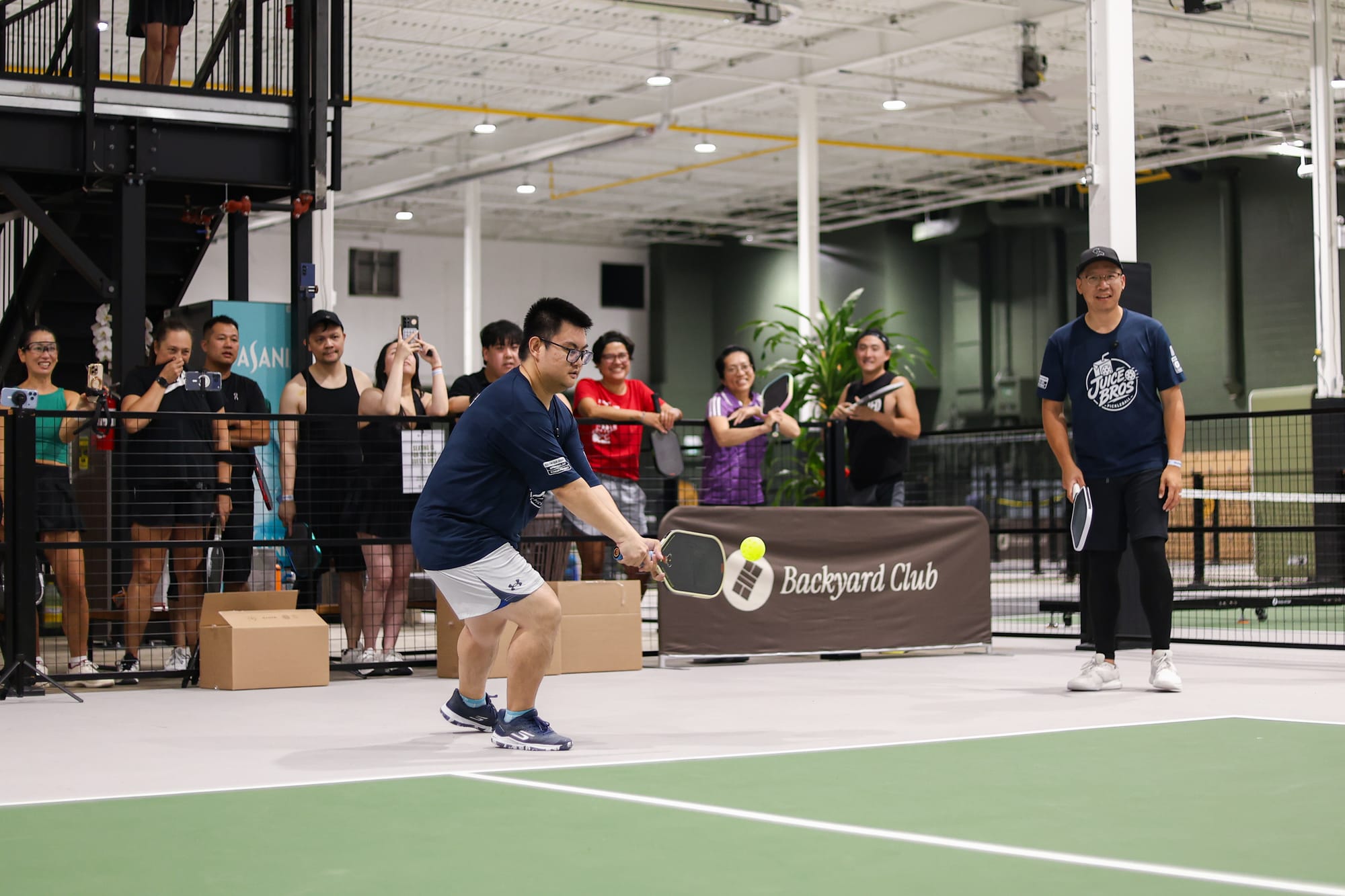Adrian reaching for a low backhand with the crowd at the rail filming, Daven waiting behind him with his paddle down